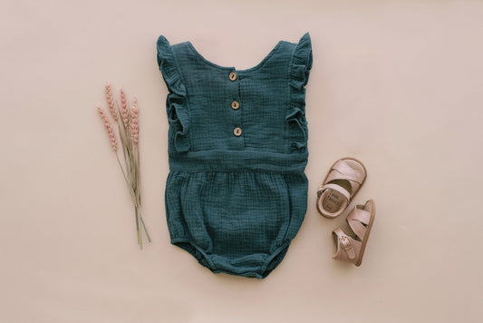 Peacock green backless baby romper with flutter sleeves, styled flat with dried pink grasses and blush leather baby sandals - Babe Basics