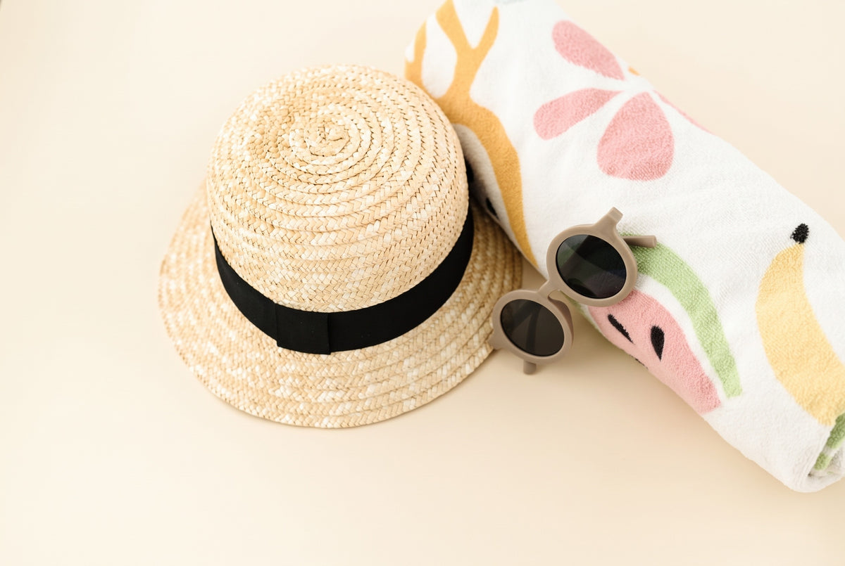 Unisex straw baby sun hat with a black band, styled with taupe toddler sunglasses and a fruit-print beach towel - Babe Basics